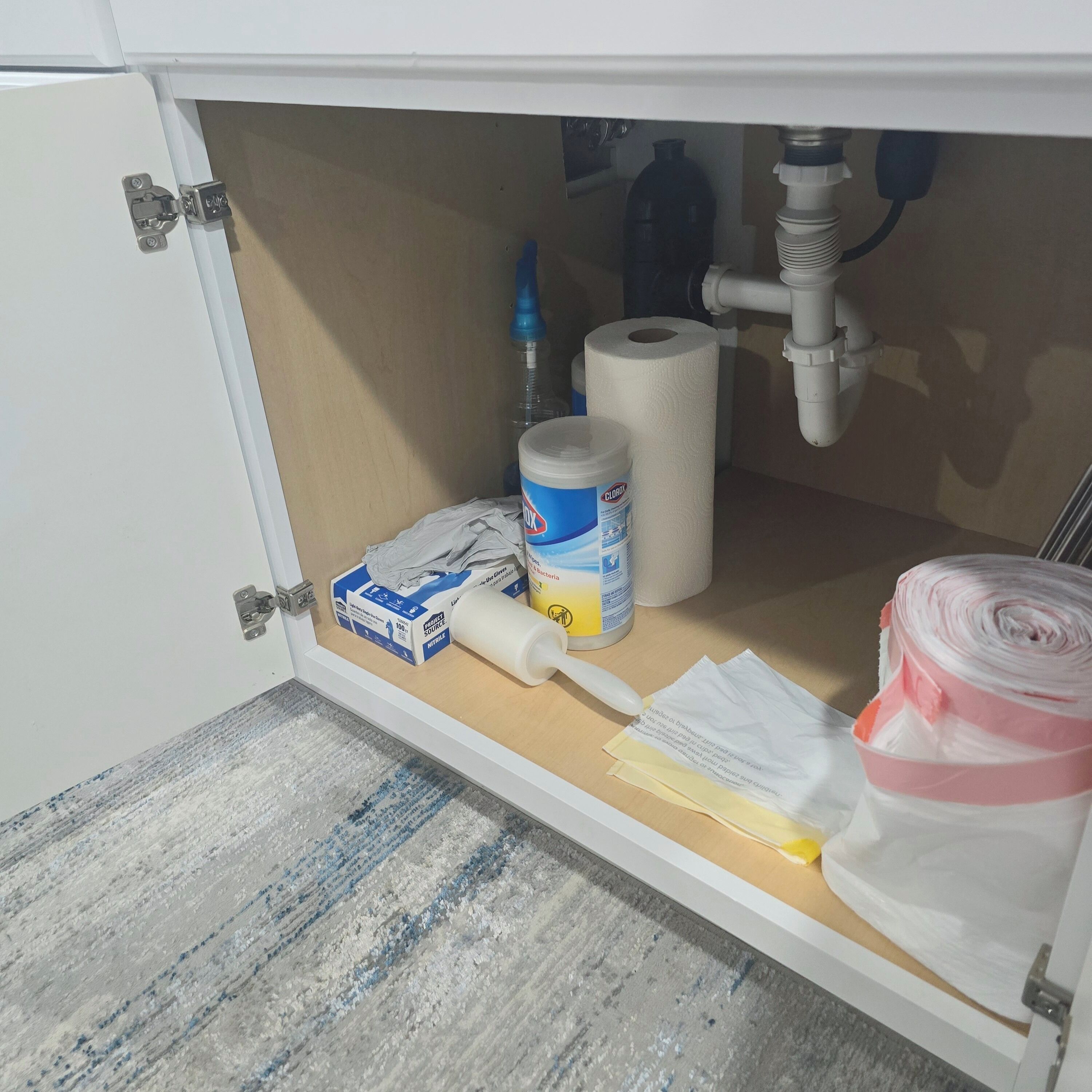 Un armario de cocina con interior blanco, que contiene un rollo de papel de cocina, un envase de limpiador Clorox, un rollo de papel higiénico y un cepillo amarillo para limpieza.