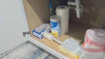Un armario de cocina con interior blanco, que contiene un rollo de papel de cocina, un envase de limpiador Clorox, un rollo de papel higiénico y un cepillo amarillo para limpieza.