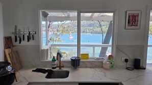 A kitchen with a large window overlooking a body of water, a sink, and various kitchen utensils and appliances. - Island escape with private waterfront beach and jetty on Scotland Island. (Scotland Island)
