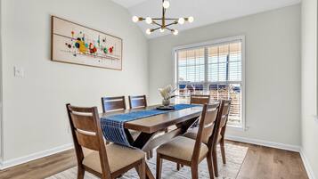 Un coin repas comprenant une table en bois, des chaises, un lustre et une fenêtre avec des stores.