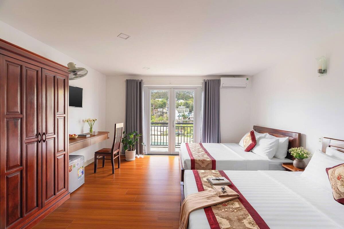 Habitación de hotel con dos camas, un armario de madera, un escritorio con silla, una planta en maceta y un balcón con cortinas.