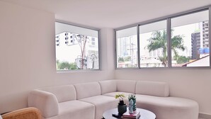 Ein modernes Wohnzimmer mit einem weißen Ecksofa, einem runden Couchtisch mit Dekoration und großen Fenstern mit Blick auf die Stadtlandschaft.