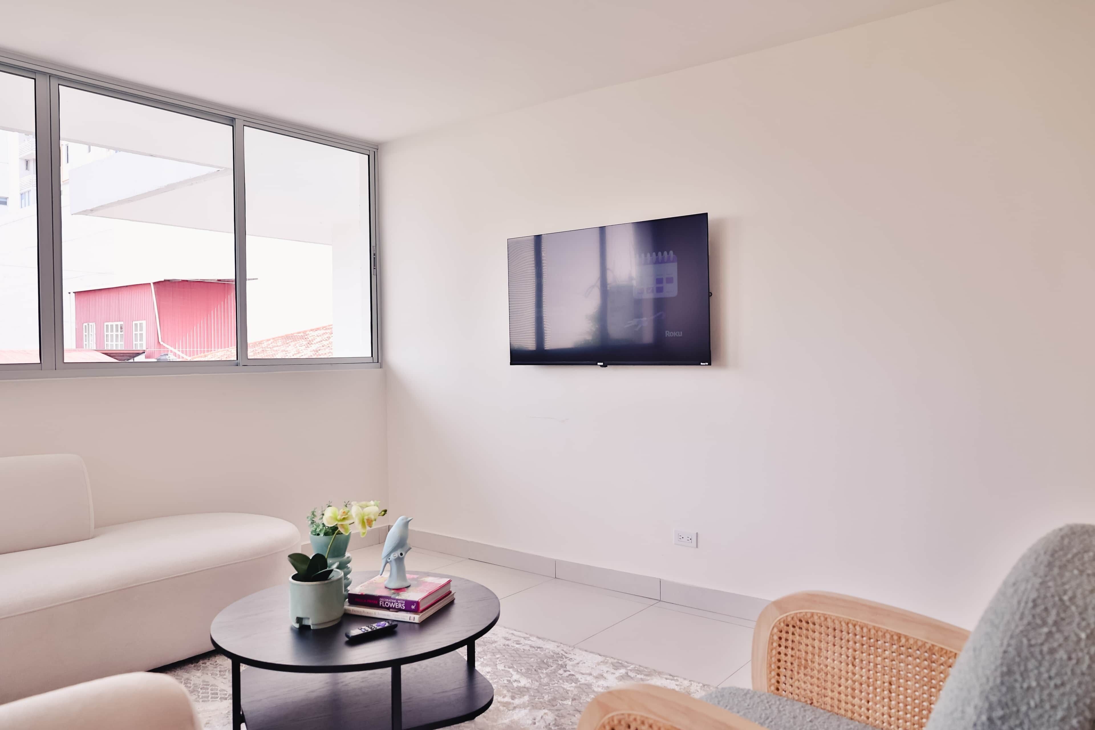 A modern living room with a white sofa, a round coffee table with decorative items, a wicker chair, and a large window with a view of a red building.