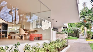 Um edifício moderno com grandes janelas de vidro, área de refeições com mesa e cadeiras, e um jardim bem cuidado com diversas plantas.