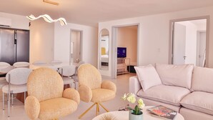 Uma sala de estar moderna com um sofá bege, duas poltronas, um espaço de jantar com mesa e cadeiras, e uma cozinha ao fundo.