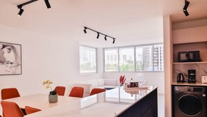 Et moderne køkken med en central ø, et spiseområde med orange stole, og et stort vindue med udsigt over byen.