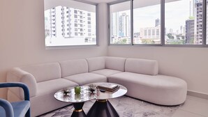 Un salon moderne avec un canapé d’angle beige, une table basse ronde en verre et un fauteuil bleu. De grandes fenêtres offrent une vue sur de hauts immeubles.