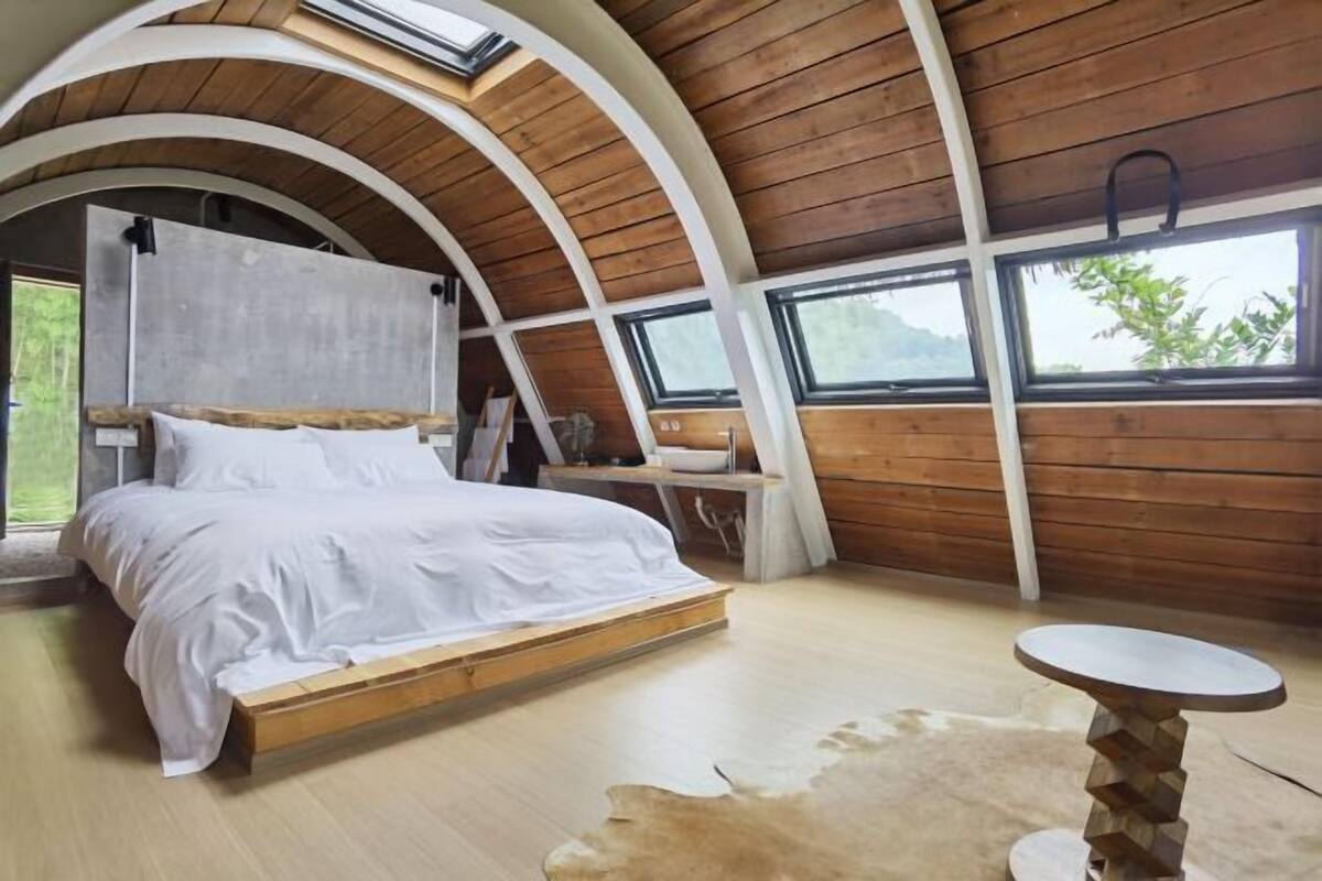 A bedroom with a wooden ceiling, a bed, a small table, and a view of the outdoors.