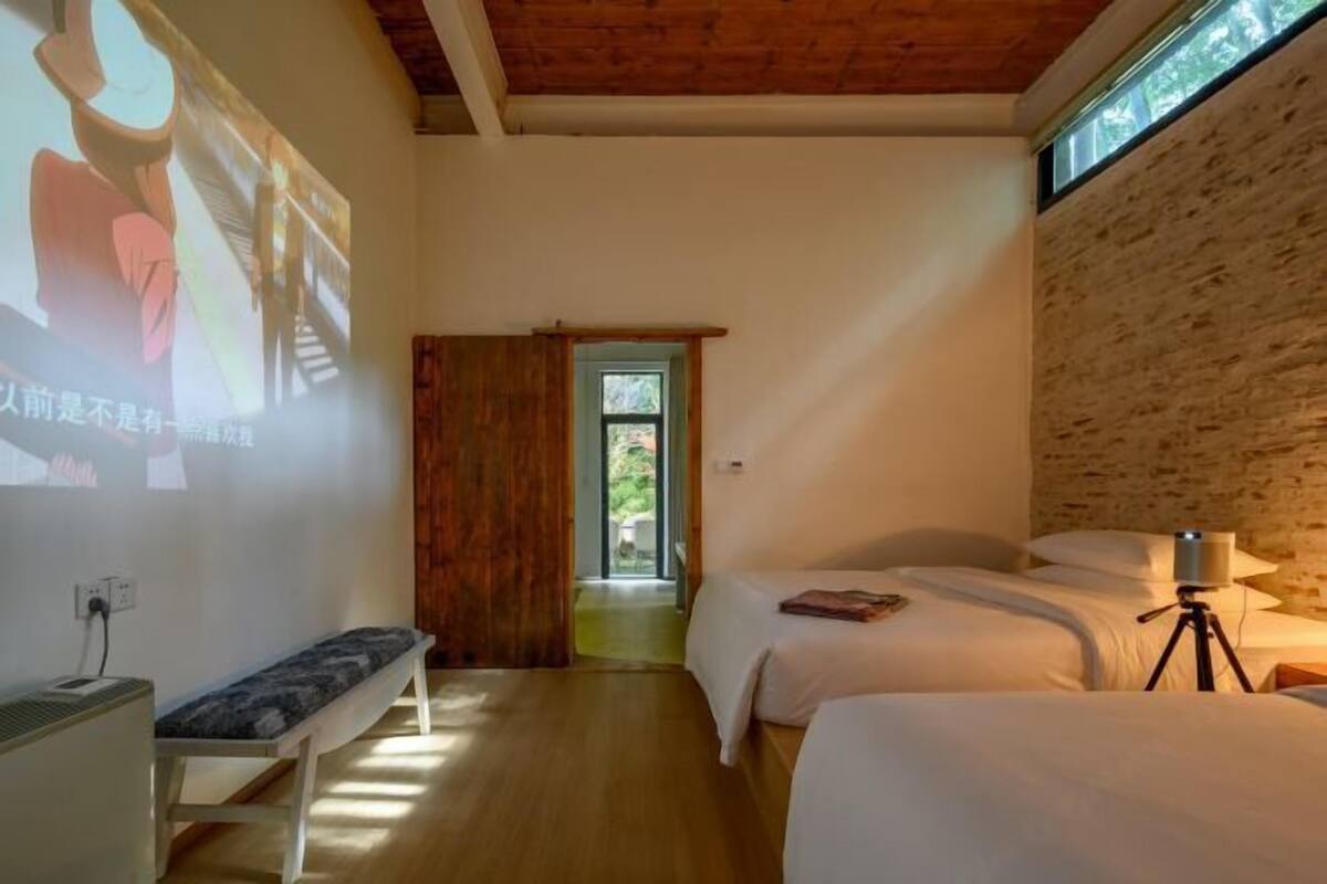 A room with a wooden ceiling, a bench, two beds, a projector screen, and a lamp.
