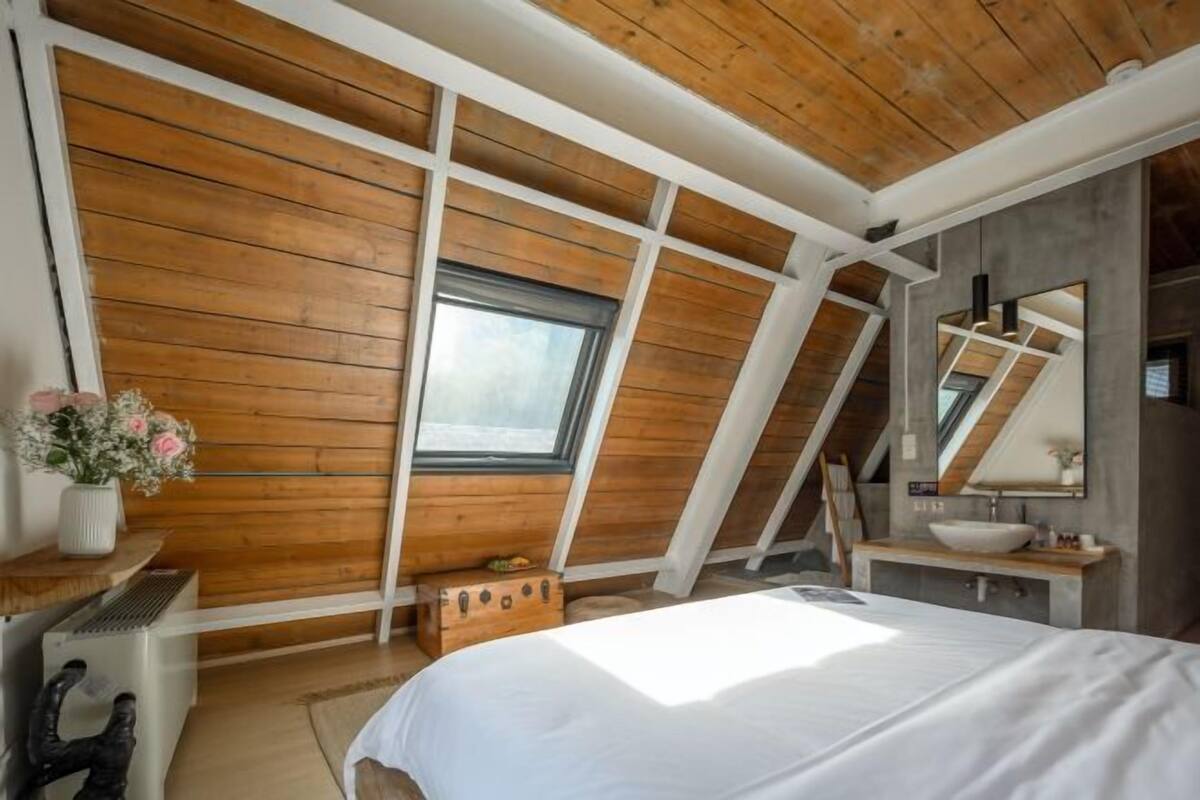 A bedroom with a sloped ceiling, wooden panel walls, a large bed, a window, a mirror, and a sink.