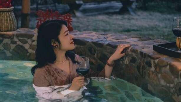 Une personne assise dans un bain à remous dont les parois sont recouvertes de pierres, tenant un verre de vin rouge.