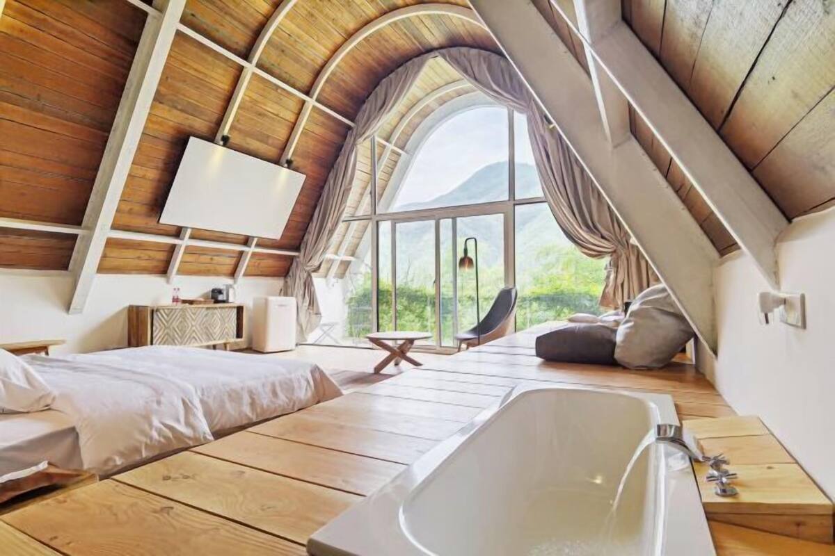 A bedroom with a sloped ceiling, a large window, a bathtub, and a bed.