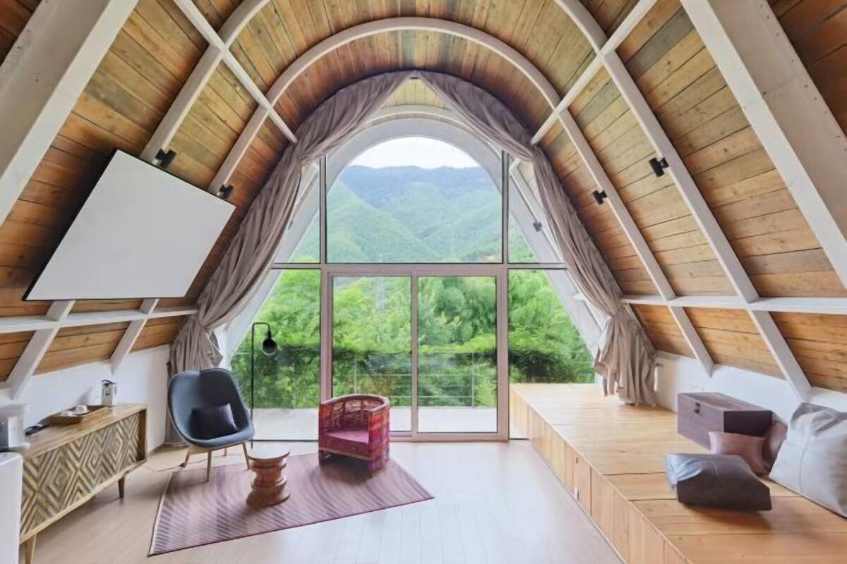 An attic room with a large window, wooden beams, a chair, a small table, and a sofa.