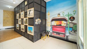 Un hall d’hôtel au design moderne, agrémenté d’une grande fresque représentant un paysage urbain, d’une cloison noire ornée de tableaux encadrés et d’un espace détente avec des tabourets.