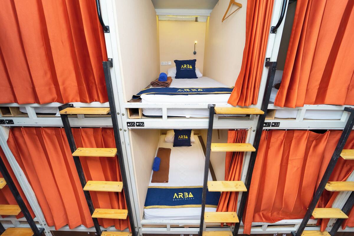 Een stapelbed met oranje gordijnen, een blauw kussen met de tekst 'ARBA' en een bruine handdoek.