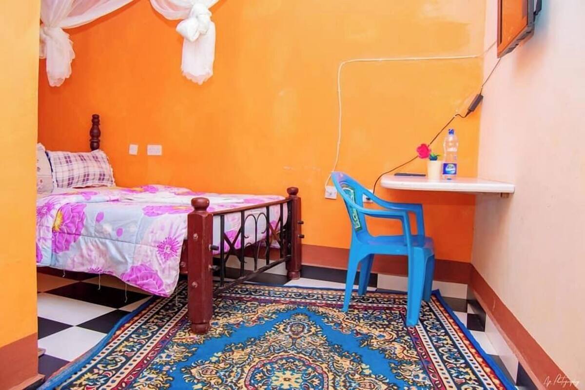 A small, brightly colored room with a bed, a blue chair, a small table, and a patterned rug.