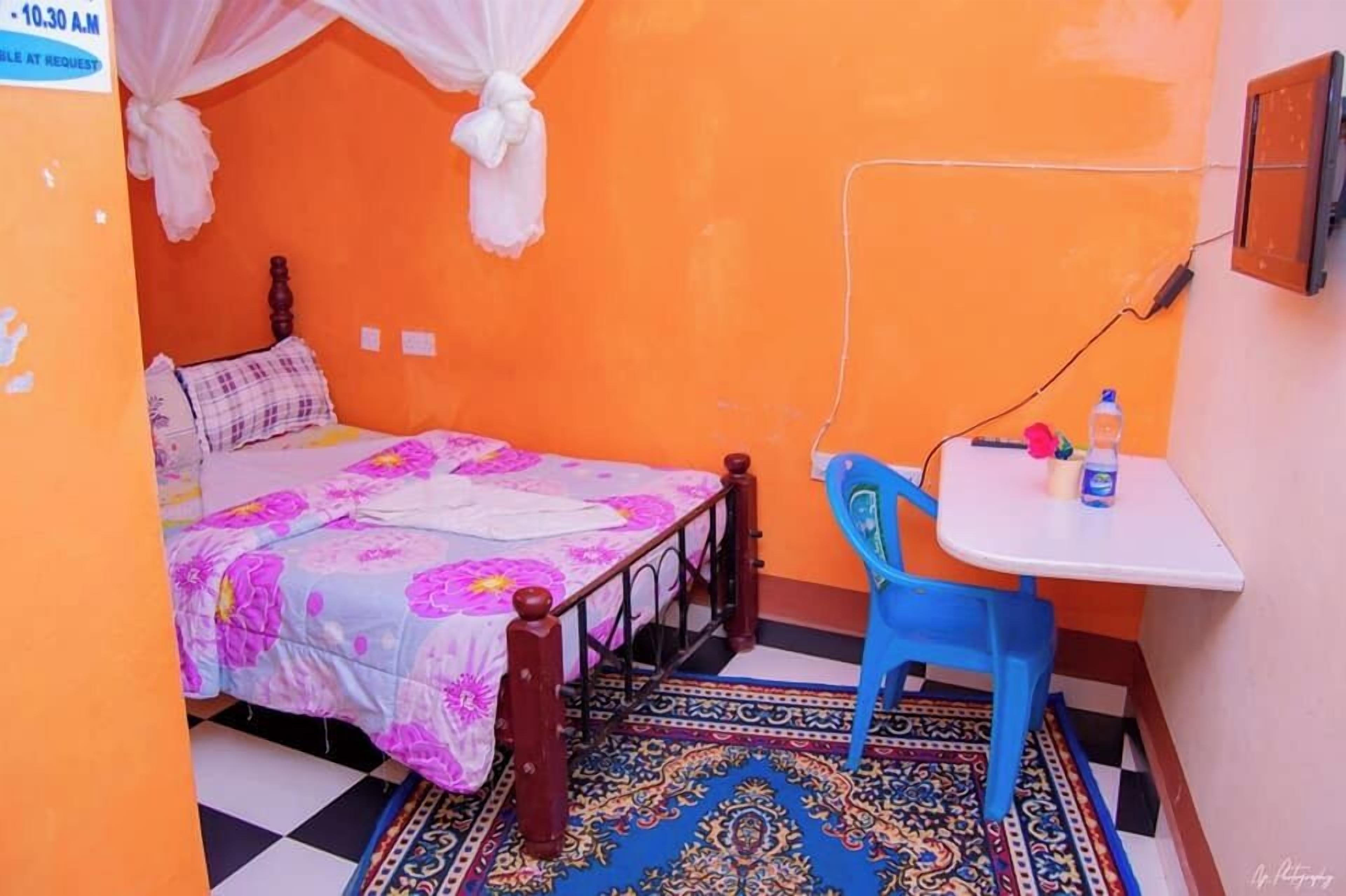 Una stanza piccola con una parete arancione, un letto con copriletto floreale, una sedia blu, un tavolino e un tappeto decorato.
