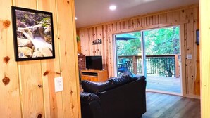 Un intérieur en bois avec une télévision, un canapé en cuir noir et une grande porte-fenêtre coulissante donnant sur une terrasse extérieure.