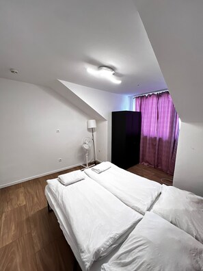 A bedroom with a sloped ceiling, a bed with white linens, a standing fan, and a window with purple curtains. - Beautiful 2BR Apt Near U6 Schoepfwerk (Vienna)