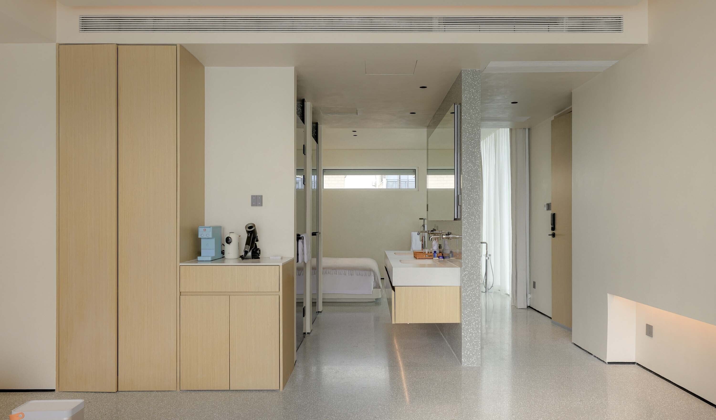 A modern hotel room with a large wardrobe, a small desk with a coffee machine, a bed, and a bathroom with a shower.