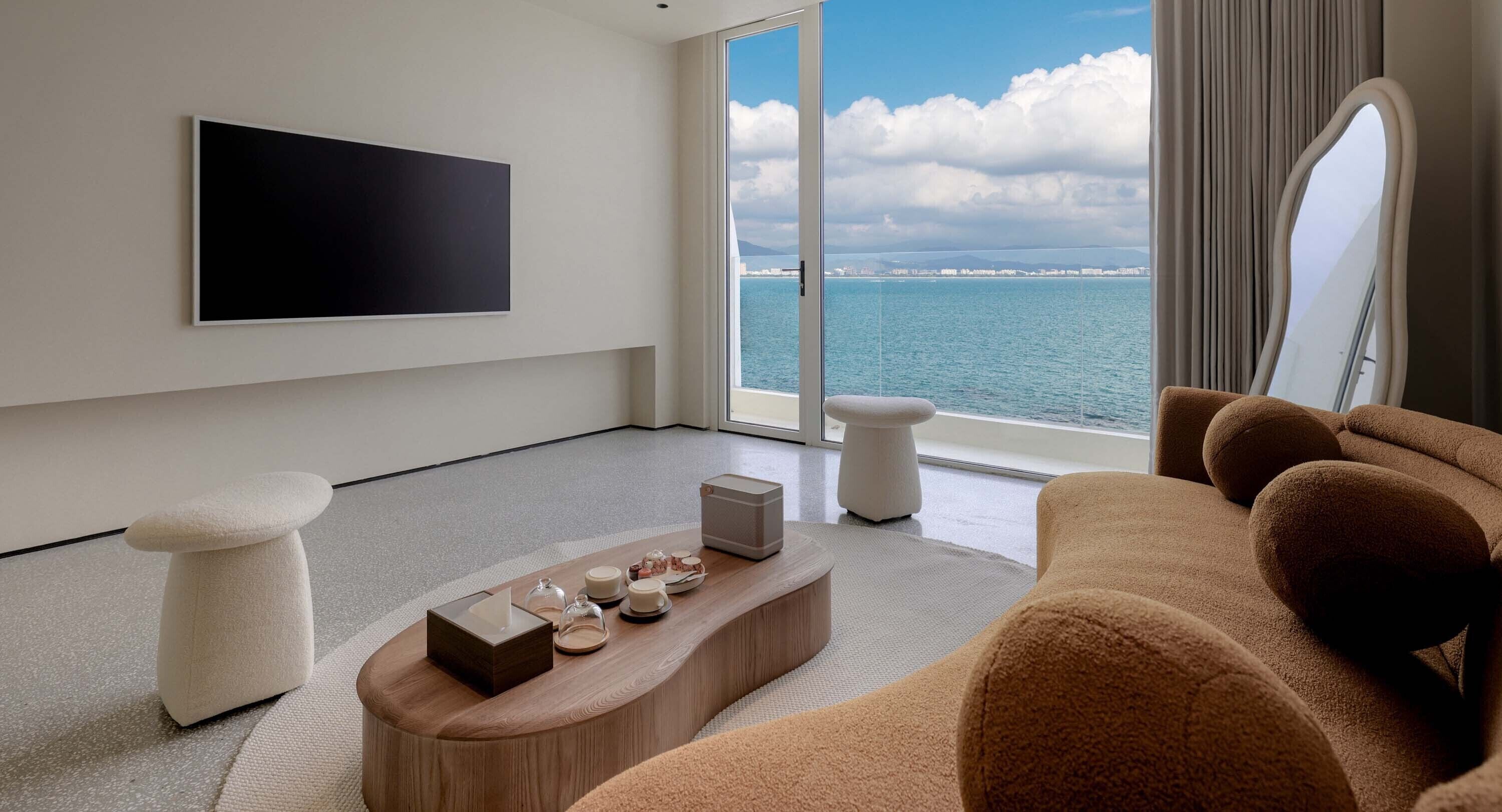 A modern living room with a large flat-screen TV, a sofa, a coffee table with decorative items, and a view of the sea through large windows.