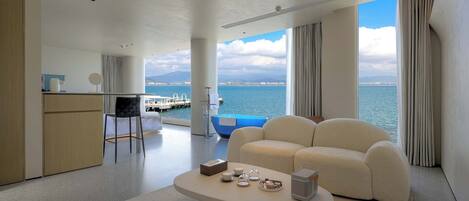 Een moderne hotelkamer met een groot raam met uitzicht op zee, een gezellige zithoek met een bijzettafeltje, en een kleine kitchenette.