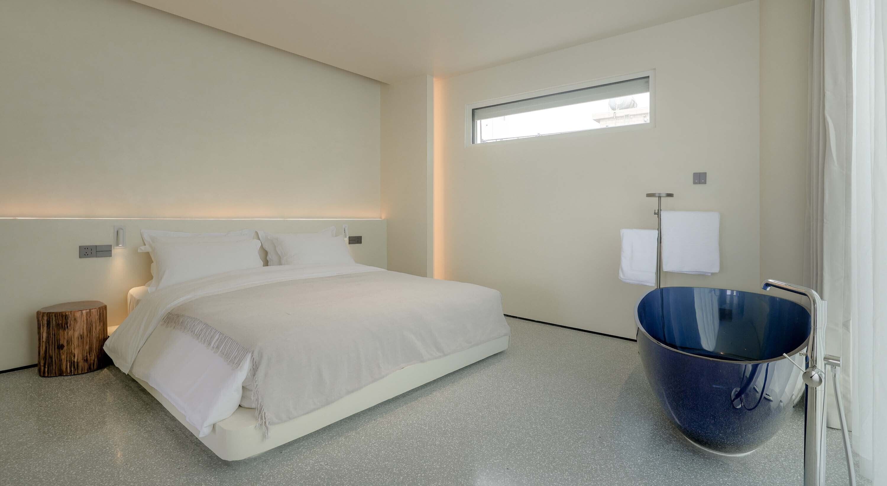 A modern hotel room with a large bed, a small bedside table, a wooden stool, a blue bathtub, and a window providing natural light.