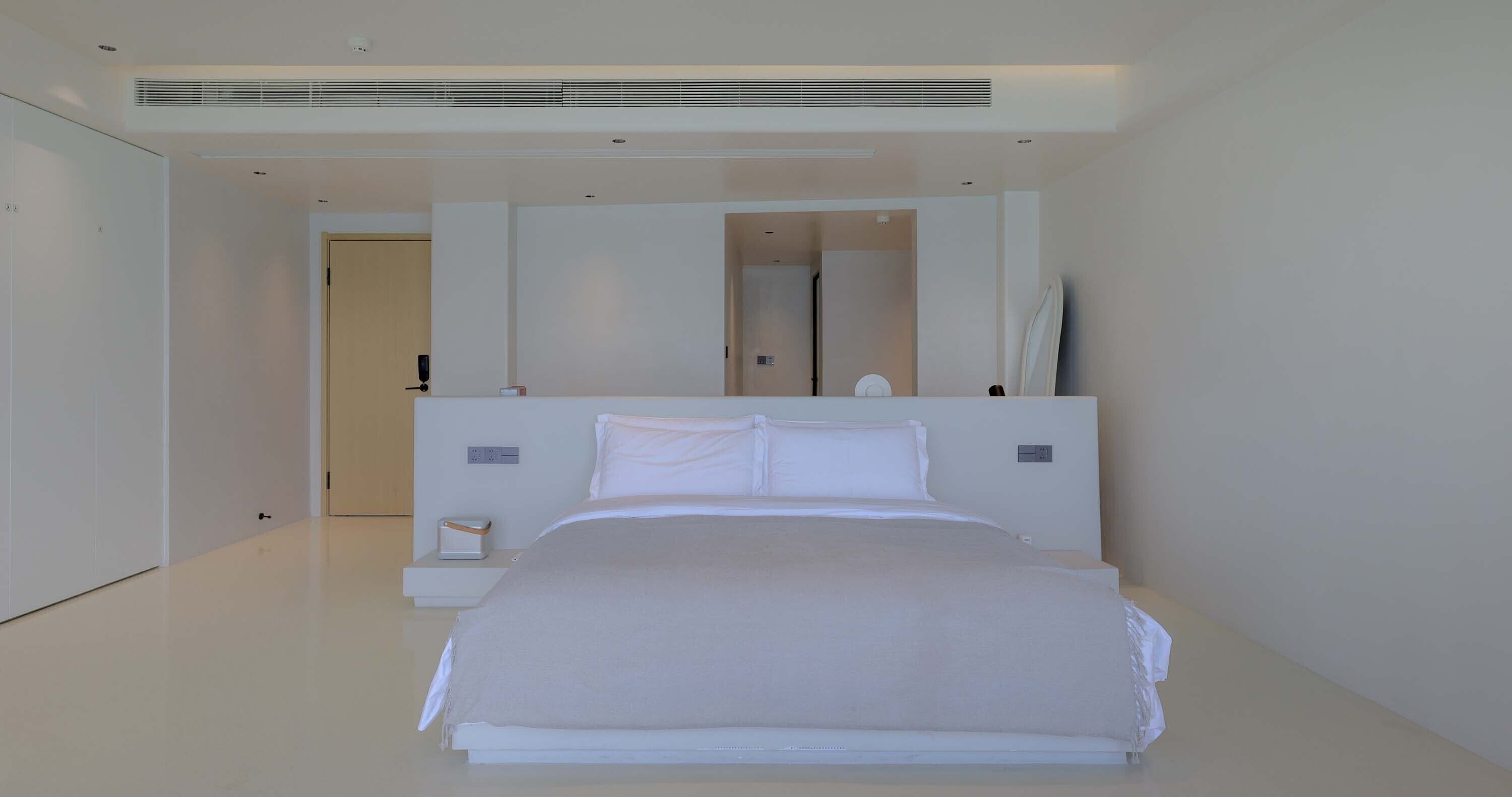 A modern hotel room with a large bed, a bedside table, a nightstand, a mirror, and a door leading to another room.