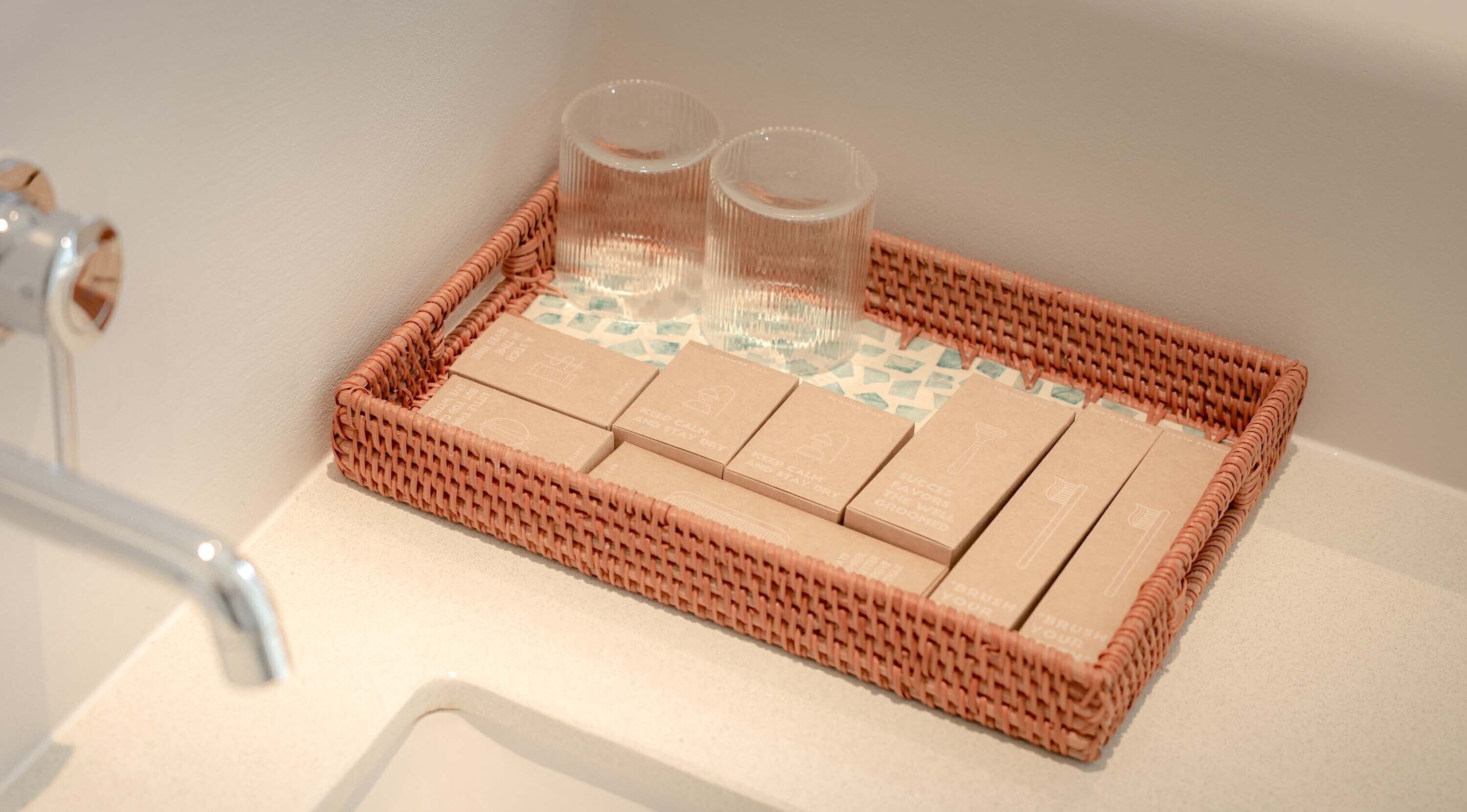 Una bandeja de mimbre con jabones y dos vasos sobre una encimera de baño.