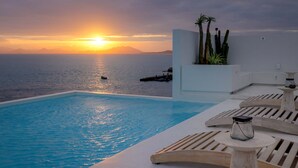 Une piscine à débordement offrant une vue sur le coucher de soleil, des chaises longues et un jardinière plantée de cactus.