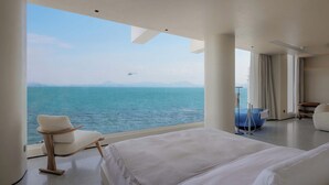 Une chambre d’hôtel moderne avec un grand lit, un coin salon avec un fauteuil, une baignoire bleue et une vue sur la mer, avec un hélicoptère visible au loin.