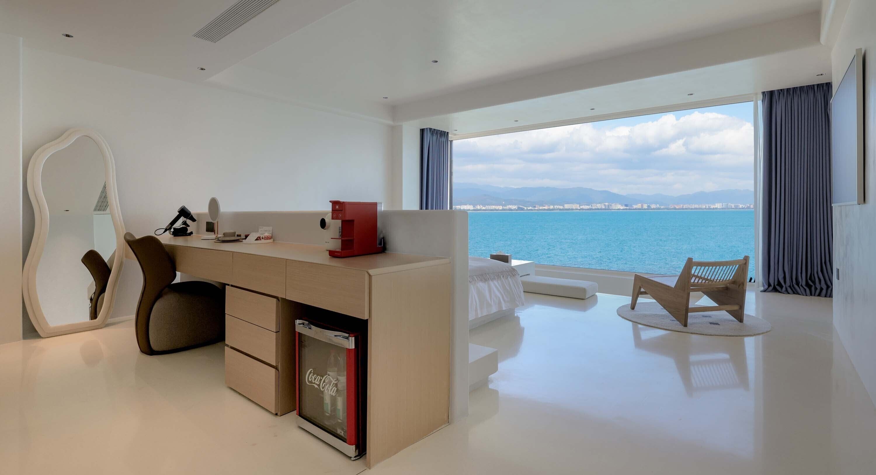 A modern hotel room with a desk, a mirror, a small fridge, a seating area with a view of the sea, and a large window with curtains.