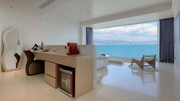 A modern hotel room with a desk, a mirror, a small fridge, a seating area with a view of the sea, and a large window with curtains.