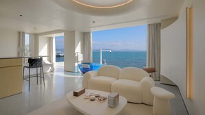 Une chambre d’hôtel moderne dotée d’une grande fenêtre donnant sur la mer, d’un coin salon avec un canapé et des fauteuils, d’un petit coin cuisine et d’une piscine à débordement bleue.