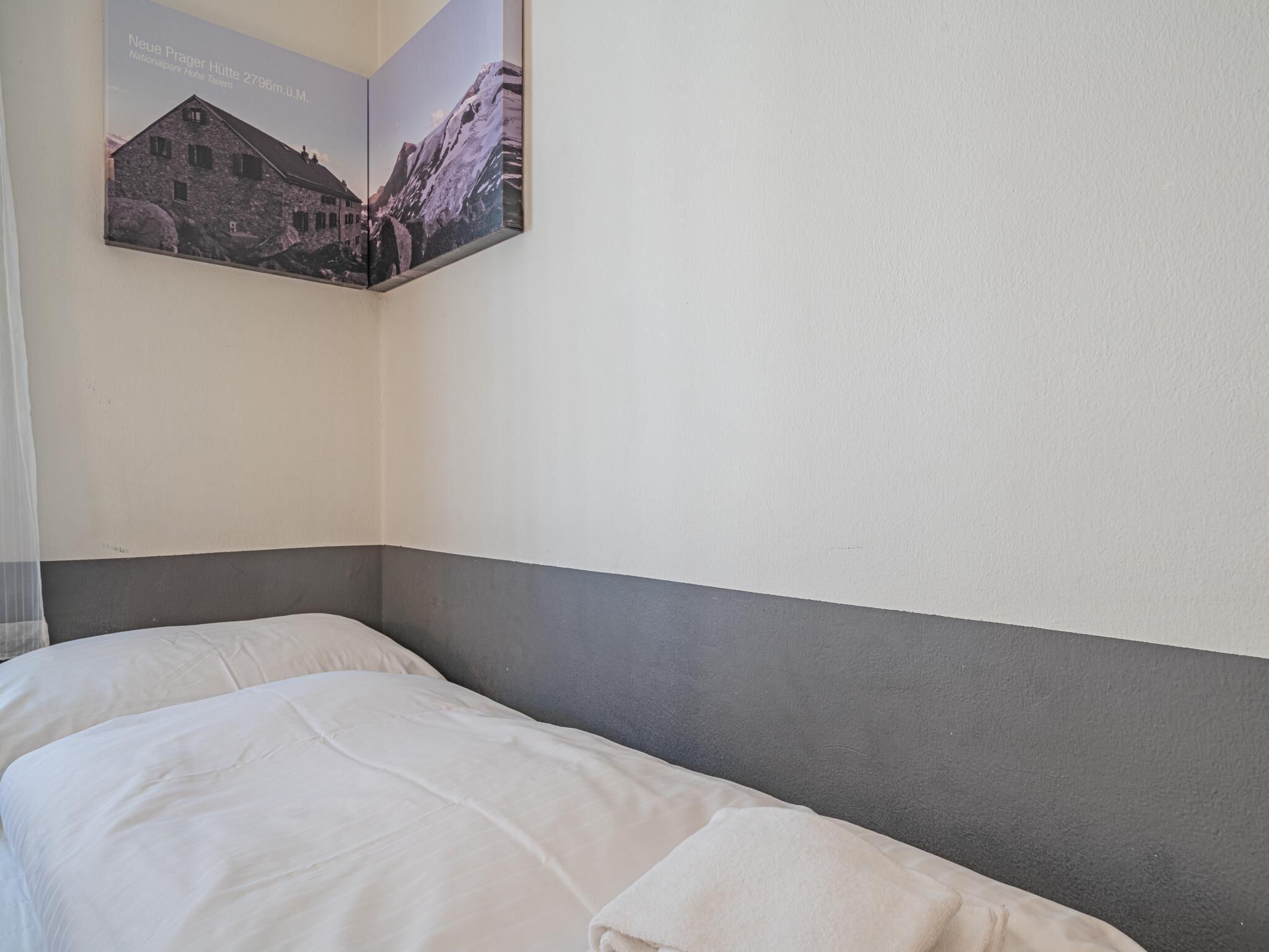 Ein Hotelzimmer mit einem Bett, einem grauen Kopfteil und einem gerahmten Bild von einem Berg und einem Gebäude an der Wand.