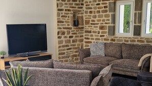 A modern living room with a stone wall, a large flat-screen TV, a wooden dining table, and comfortable seating. - The house West side, SPA, Sea at 80 meters (Plounéour-Brignogan-plages)