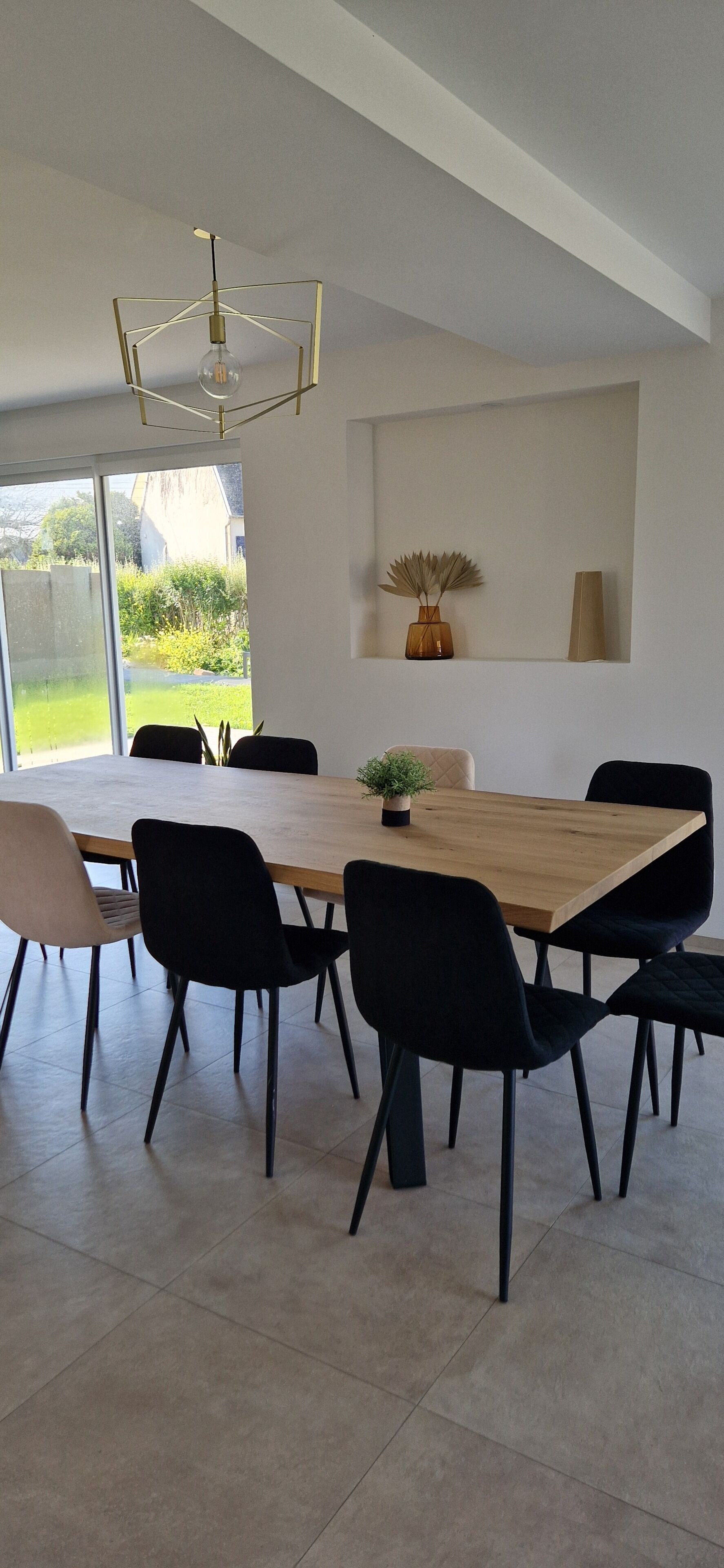 A modern dining area with a wooden table and black chairs, a pendant light, and a view of a garden through large windows.