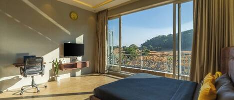 A hotel room with a large bed, a desk with a TV, and a balcony with a view of the landscape.
