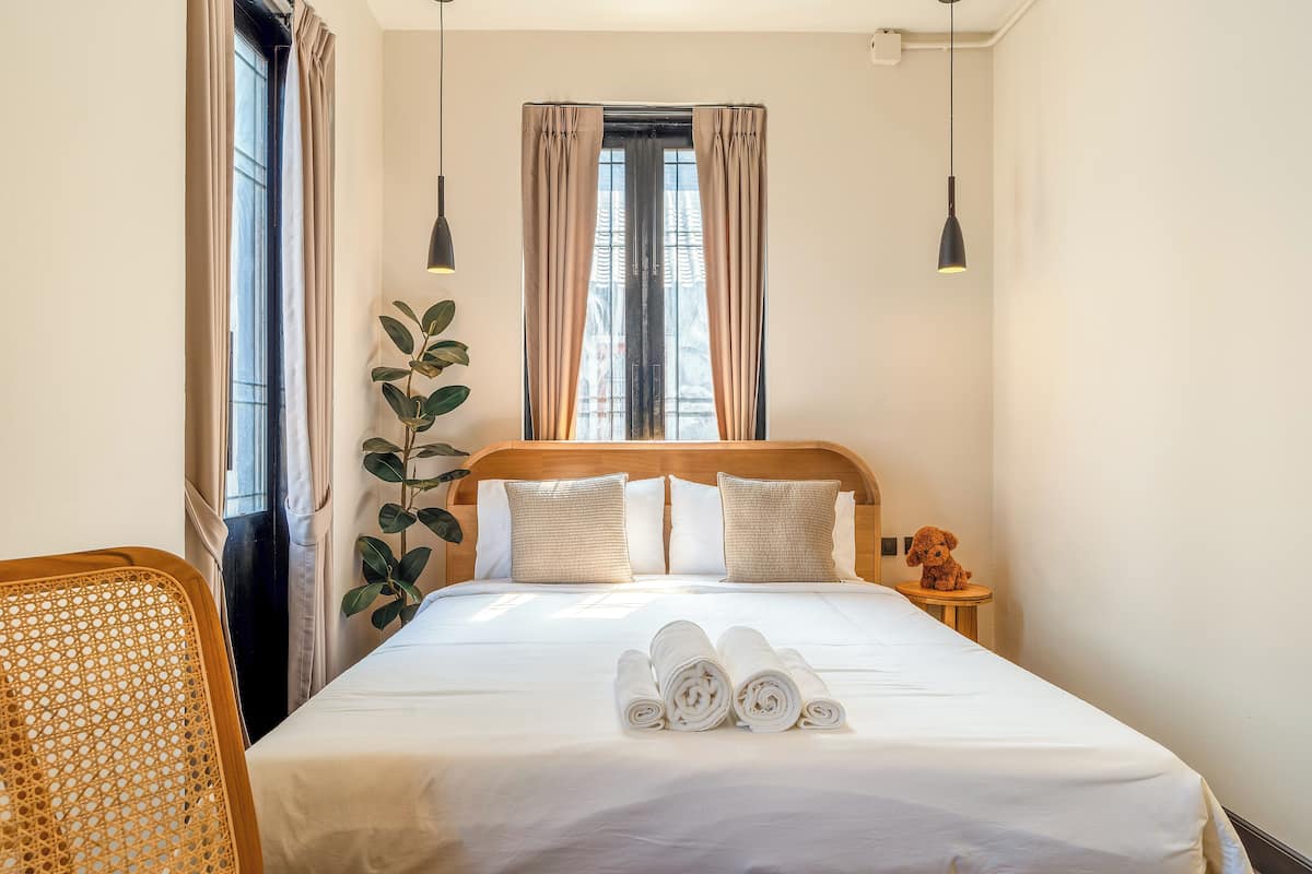 A neatly made bed with rolled towels, a wooden headboard, a bedside plant, and a teddy bear on a nightstand.