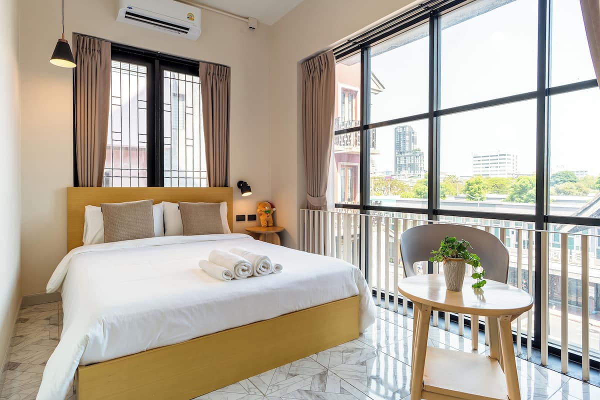 A hotel room with a large bed, a small table with a plant, and a balcony with a view of the city.