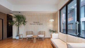 Egy modern belső tér, melynek része egy fa padló, két fonott szék, egy fehér kanapé, egy virágos növény és egy fal, melyen a „TRAVIS Samyian Bangkok” felirat látható.
