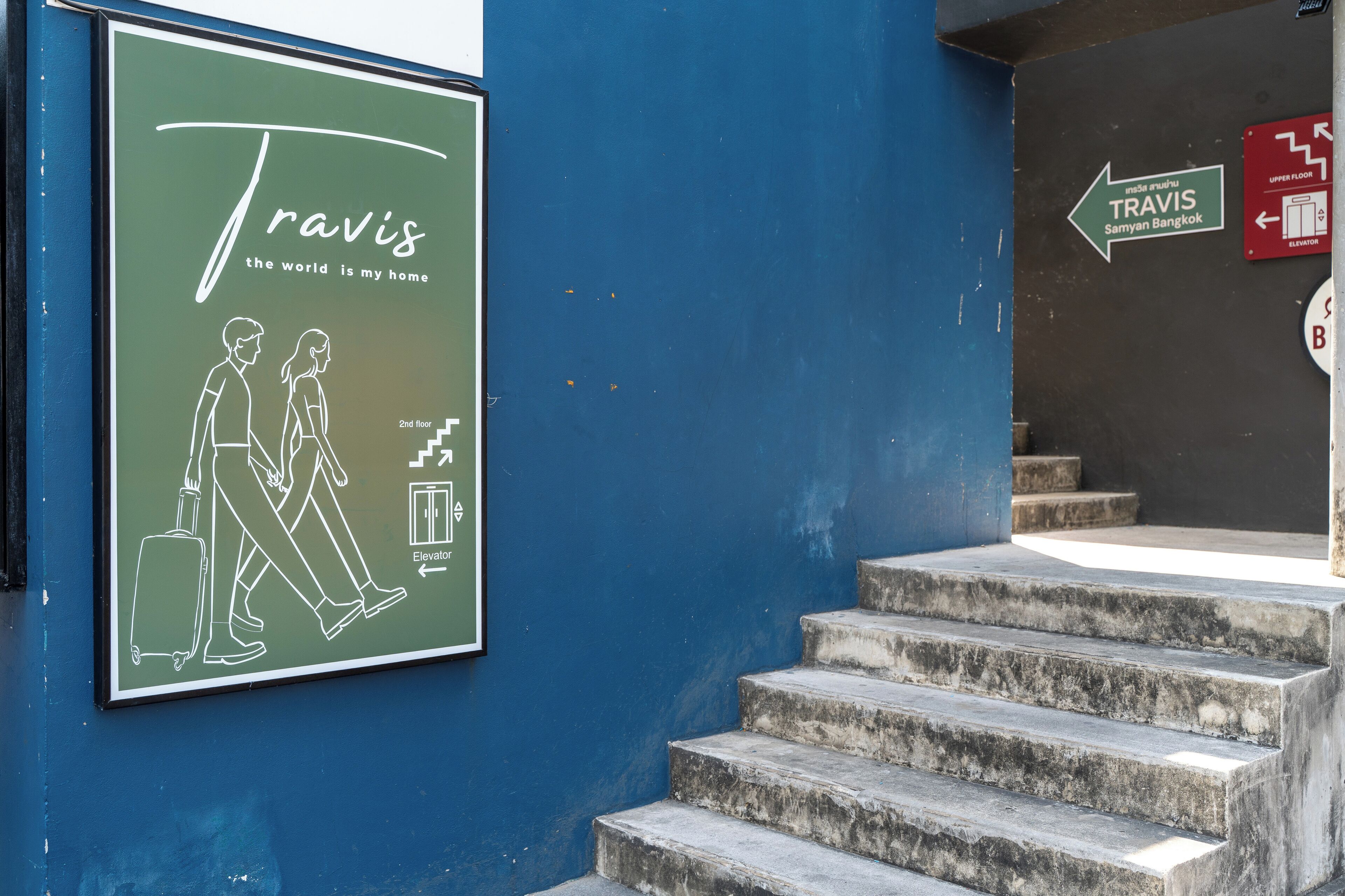 A staircase leading to an upper floor, a sign for Travis Samy Bangkok, and a poster with a walking couple and a suitcase.