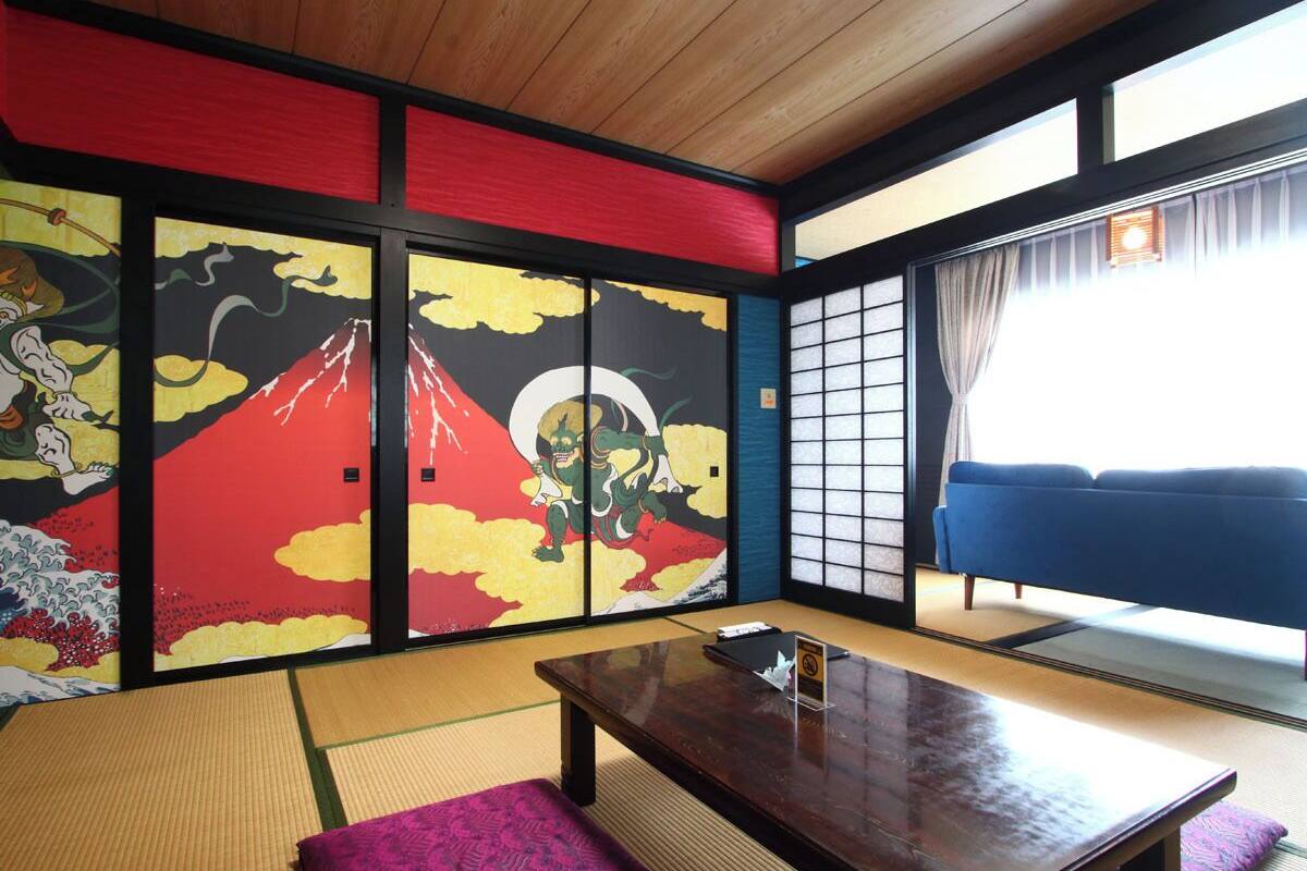 Ein traditioneller japanischer Raum mit Tatami-Boden, einem niedrigen Holztisch, einem blauen Sofa und einer Schiebetür mit farbenfrohen Bildern.