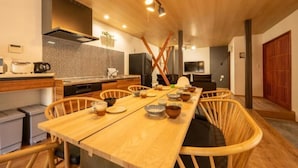 A modern kitchen with a long wooden table, wicker chairs, and various kitchen appliances. - SAKURATARO MOMIJIJIRO NO MIYAKO (Asakura)