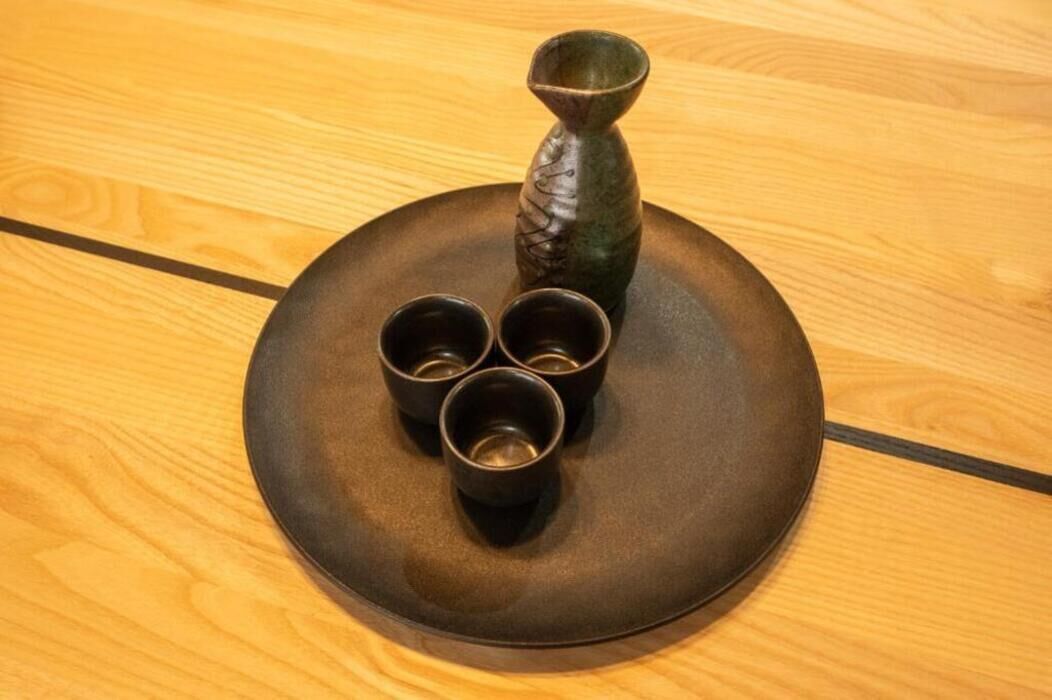 A round wooden table with a dark plate, a decorative vase, and three small cups.
