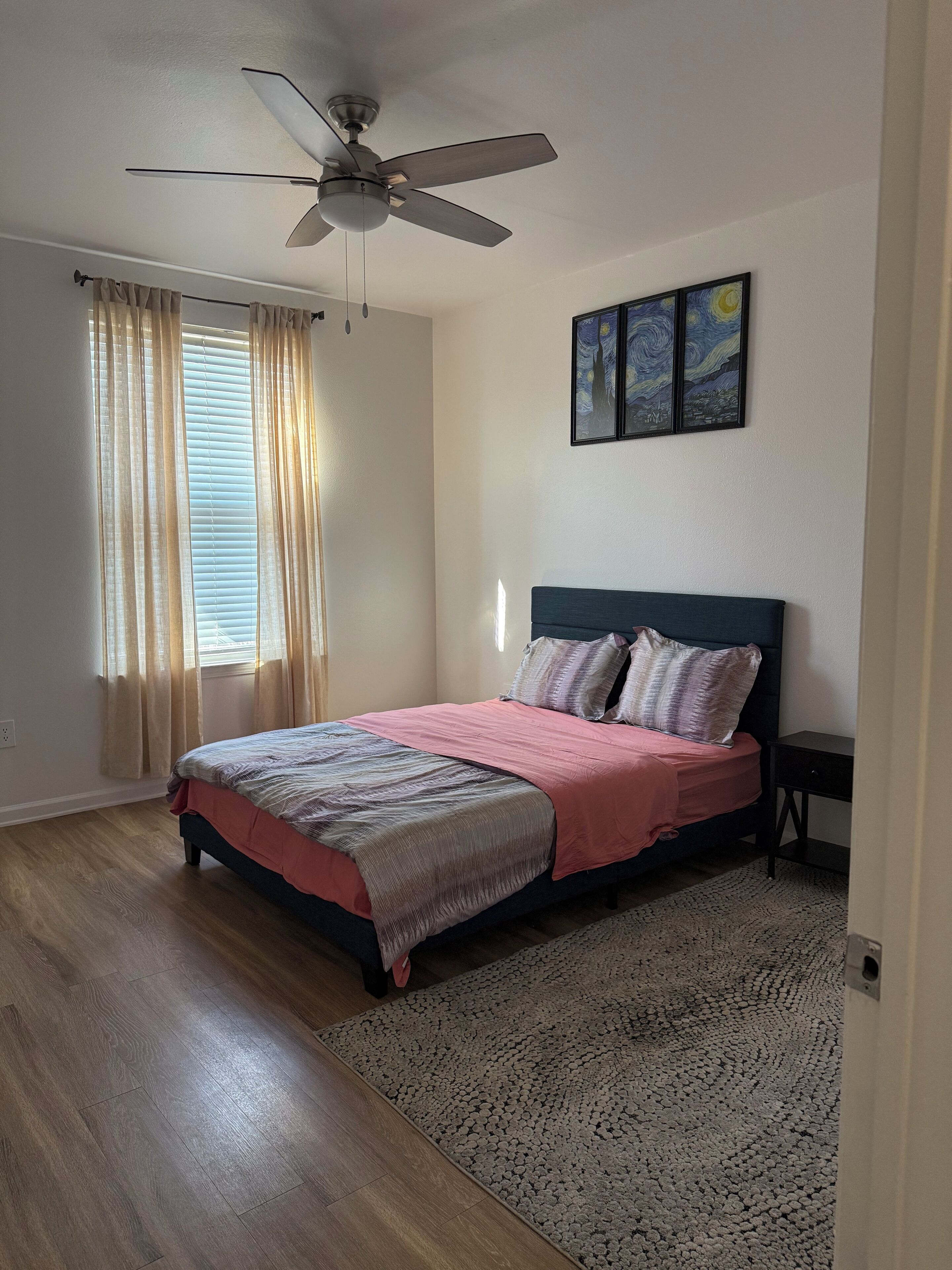 A bedroom with a bed, a ceiling fan, a window with curtains, a rug, and artwork on the wall.