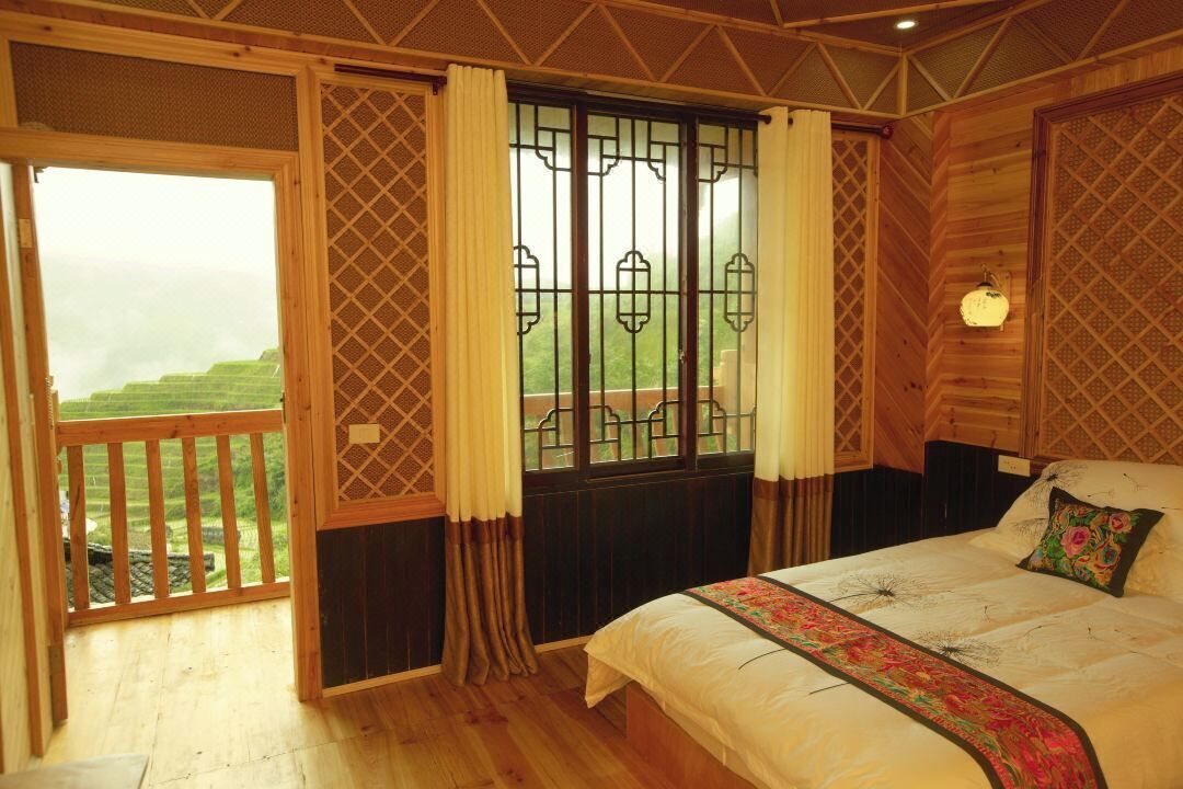 A bedroom with a large bed, a view of green fields through a window, and a balcony with wooden railings.