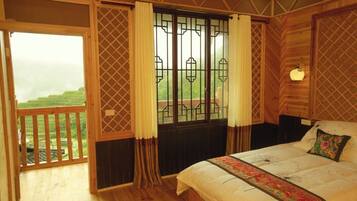A bedroom with a large bed, a view of green fields through a window, and a balcony with wooden railings.