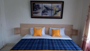 Ein Hotelzimmer mit einem Bett, zwei Kissen, einer blau-gelben Tagesdecke und einem Bild an der Wand.