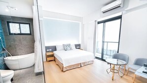 Egy modern szállodai szoba, amelyben egy nagy ágy, külön fürdőszoba, egy kis asztal található, és egy nagy ablakon keresztül nyílik kilátás.